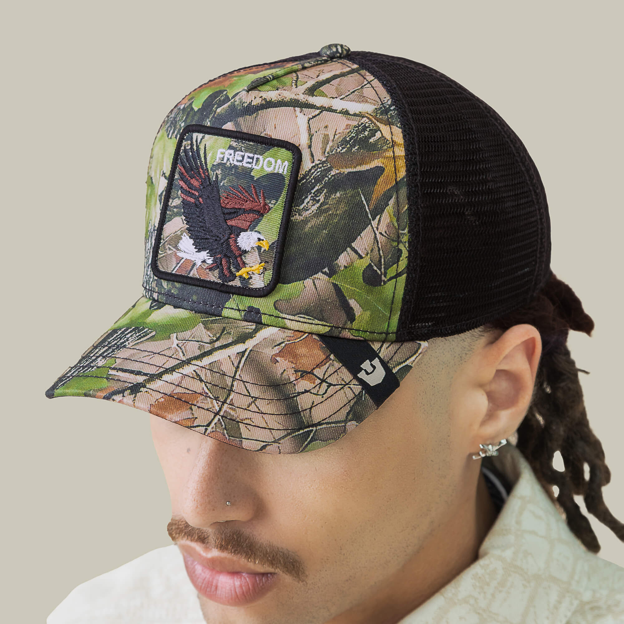 A person wears the Goorin Bros. Camouflage Eagle trucker hat with eagle embroidery and a FREEDOM patch, featuring a curved brim and black mesh back—perfect for adding bold, patriotic style to any look.