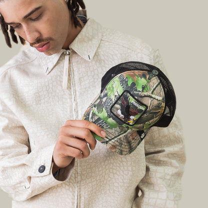 A person in a patterned beige jacket holds the Goorin Bros. Camouflage Eagle trucker hat, featuring eagle embroidery and a bold FREEDOM patch—a true statement piece.