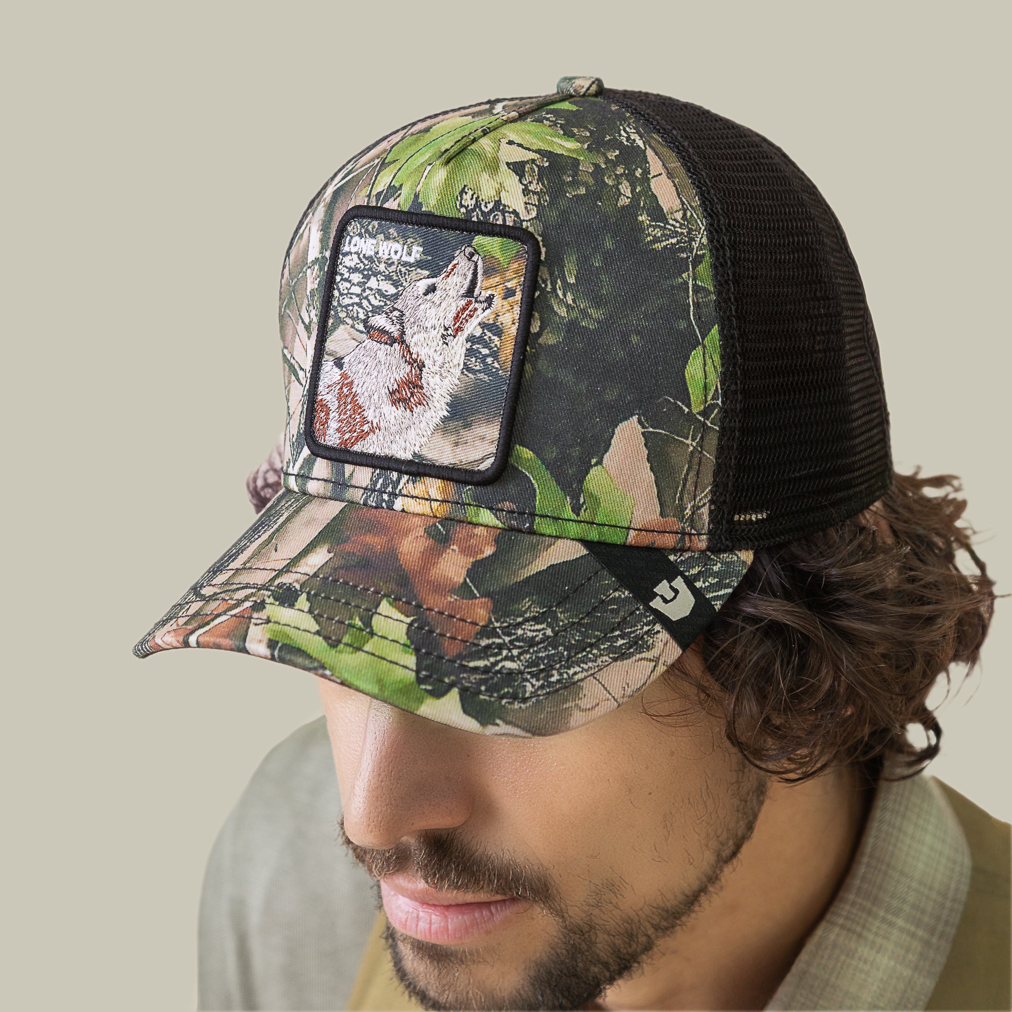A man sporting the Goorin Bros. Camouflage Lone Wolf trucker hat—featuring an embroidered howler wolf patch and bold "WOLF" lettering—a killer wardrobe choice for any off-grid soft launch.