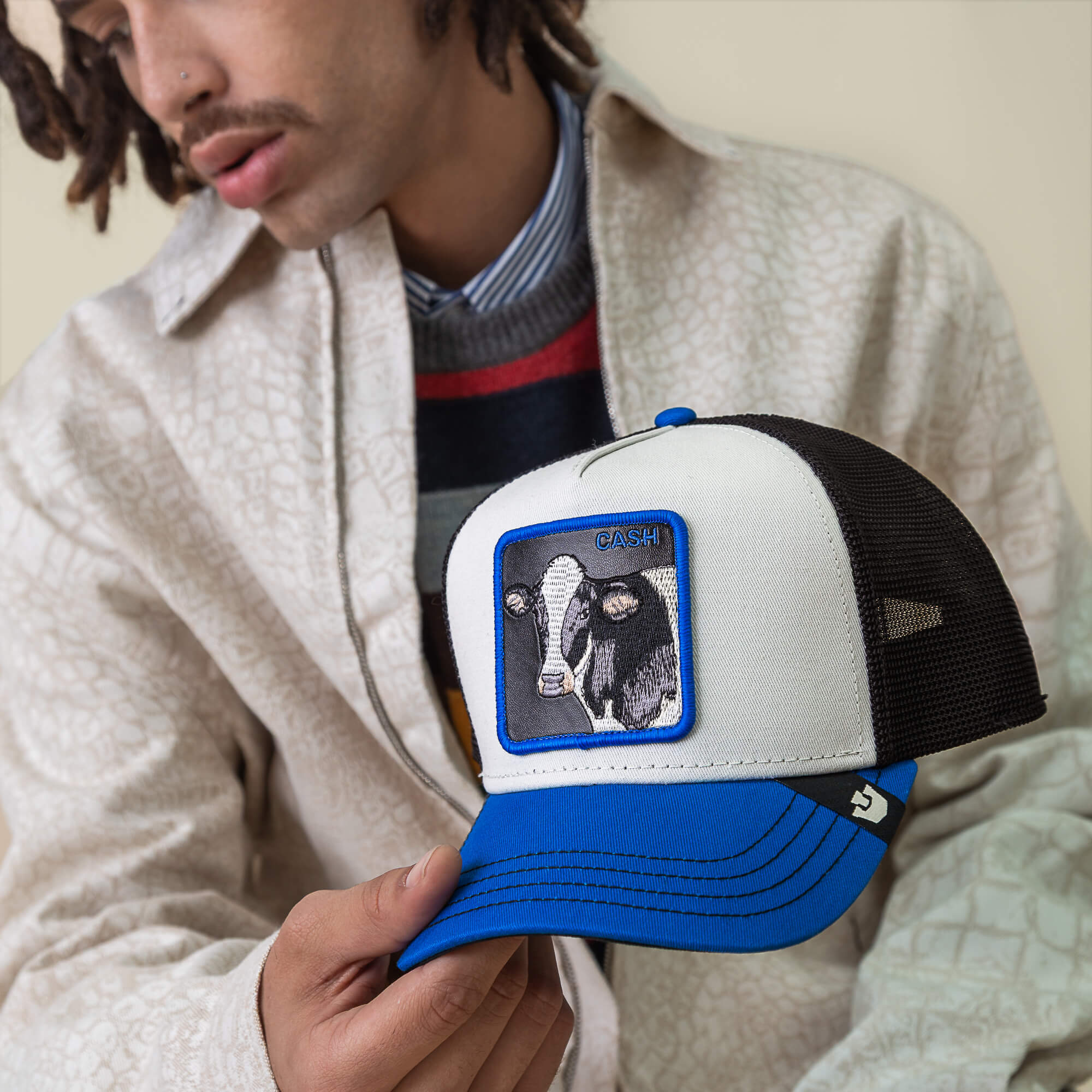 A man in a light patterned jacket holds the Goorin Bros. Tricolor Cow trucker hat, featuring a CASH cow patch on the front, paired with his bold wallet for a modern touch.