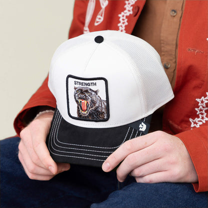 A person in a red jacket holds the Goorin Bros. “Suede Panther” cap, featuring a white crown, black brim, and a panther patch labeled "STRENGTH.