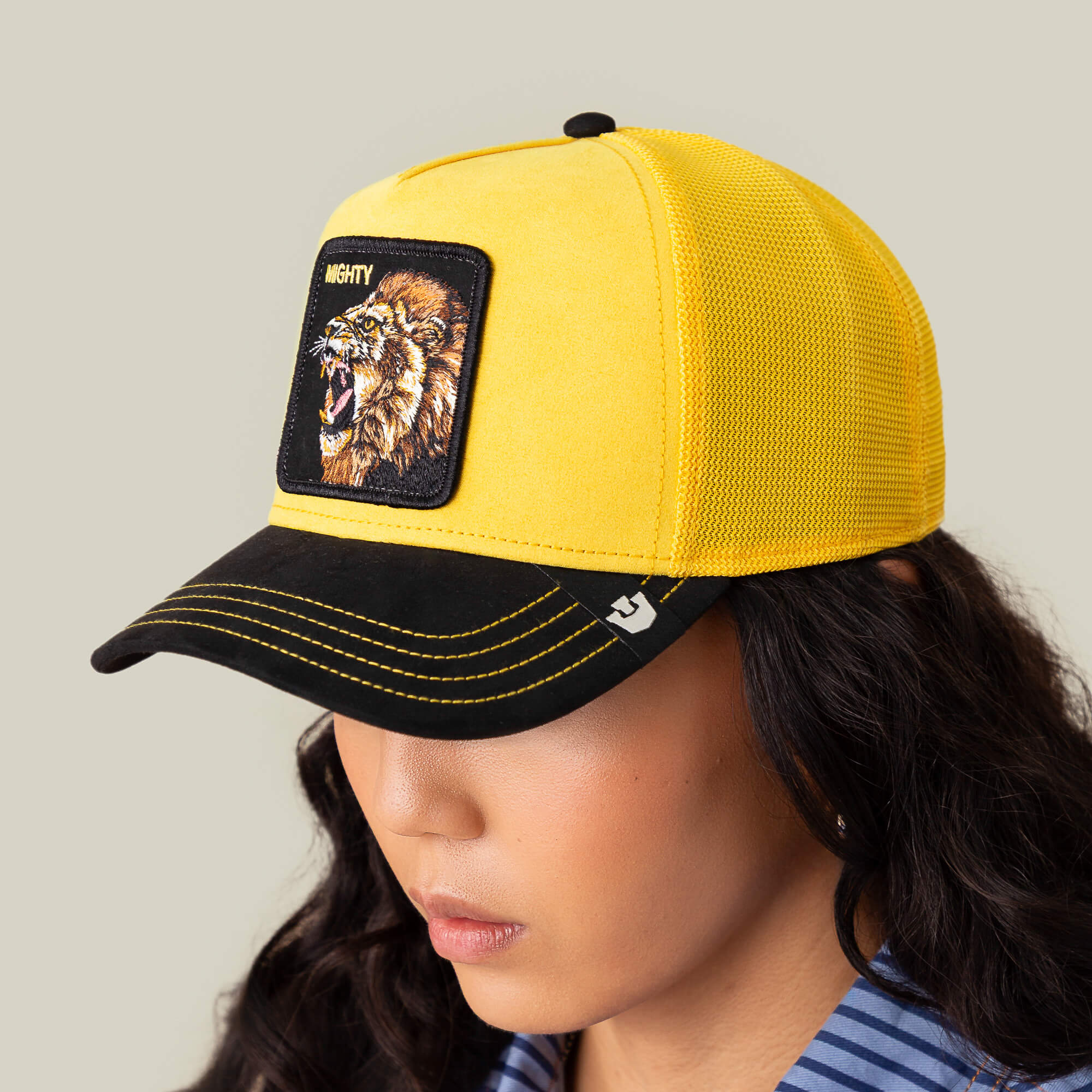 A person wearing the Goorin Bros. Suede Lion yellow and black trucker cap with a lion patch labeled MIGHTY, looking down against a plain beige background.