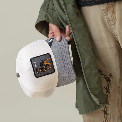 Someone in tan pants and a green jacket holds two Goorin Bros. caps by their brims: one clean white with a patch, and the other gray—the Denim Bad Boy by Goorin Bros.
