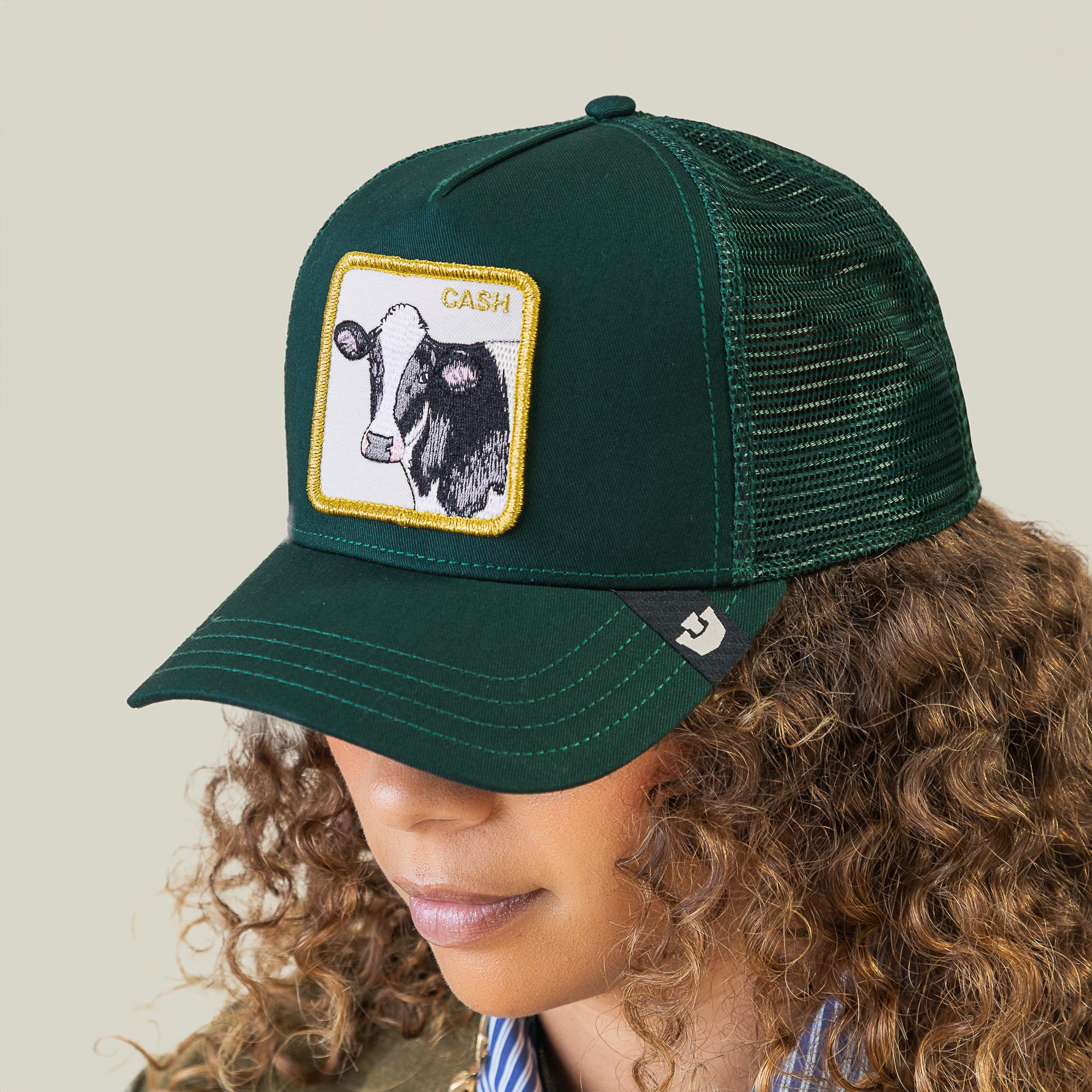A person wears the Goorin Bros. Cash Cow Trucker, a dark green mesh cap featuring a patch with a bold cow graphic and “Cash Cow” text.