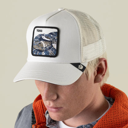 A person wearing the Goorin Bros. Toxic Trucker hat—a white trucker cap with an adjustable mesh back, decorated with a rattlesnake patch and the word “TOXIC”—paired with a gray jacket featuring an orange hood.