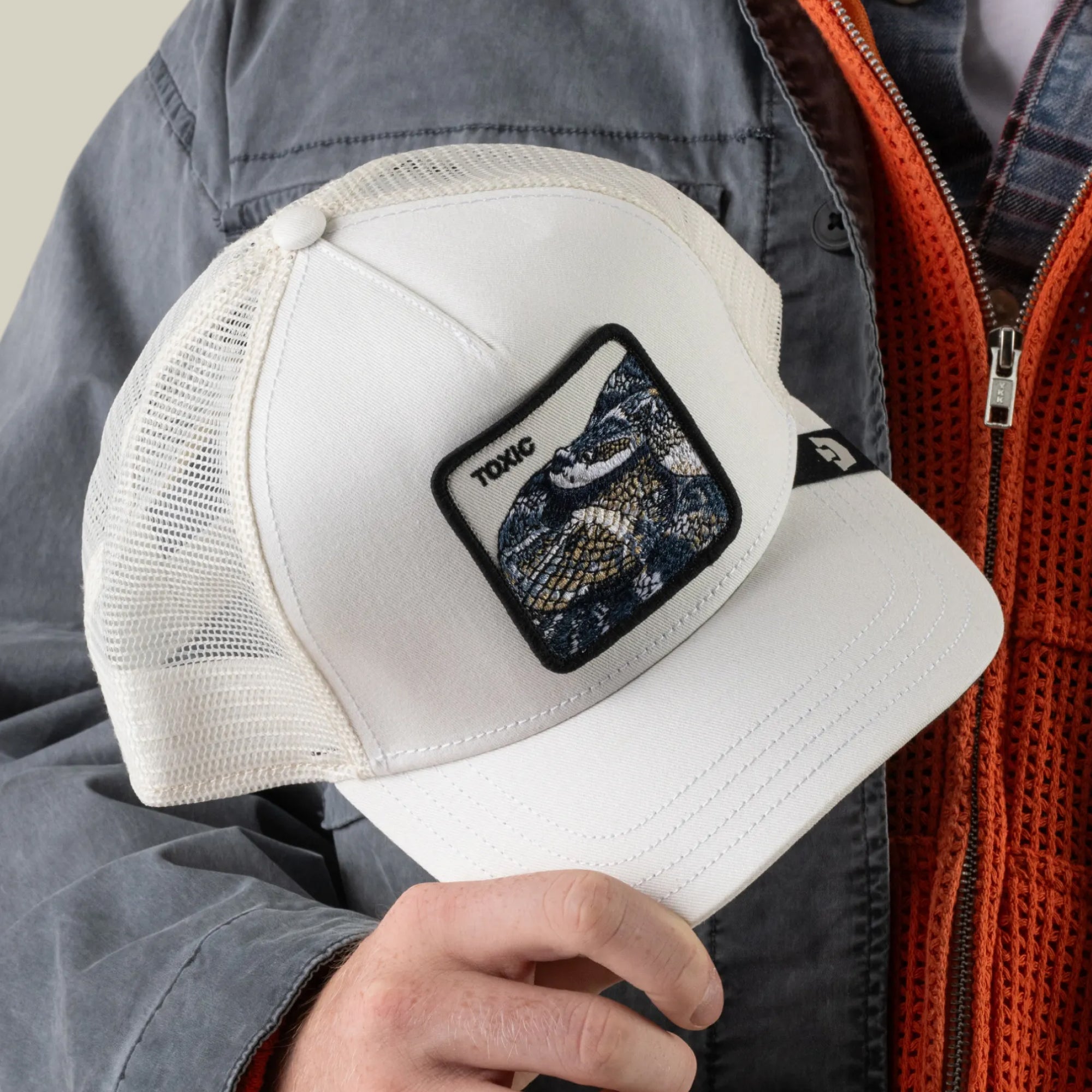 A person wearing a gray jacket holds the Goorin Bros. Toxic Trucker hat, a white trucker with an adjustable mesh back featuring a rattlesnake patch and the word “TOXIC.”.