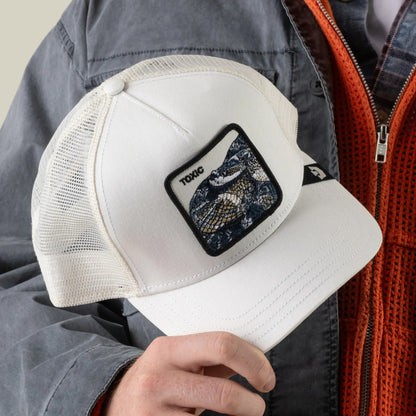 A person wearing a gray jacket holds the Goorin Bros. Toxic Trucker hat, a white trucker with an adjustable mesh back featuring a rattlesnake patch and the word “TOXIC.”.
