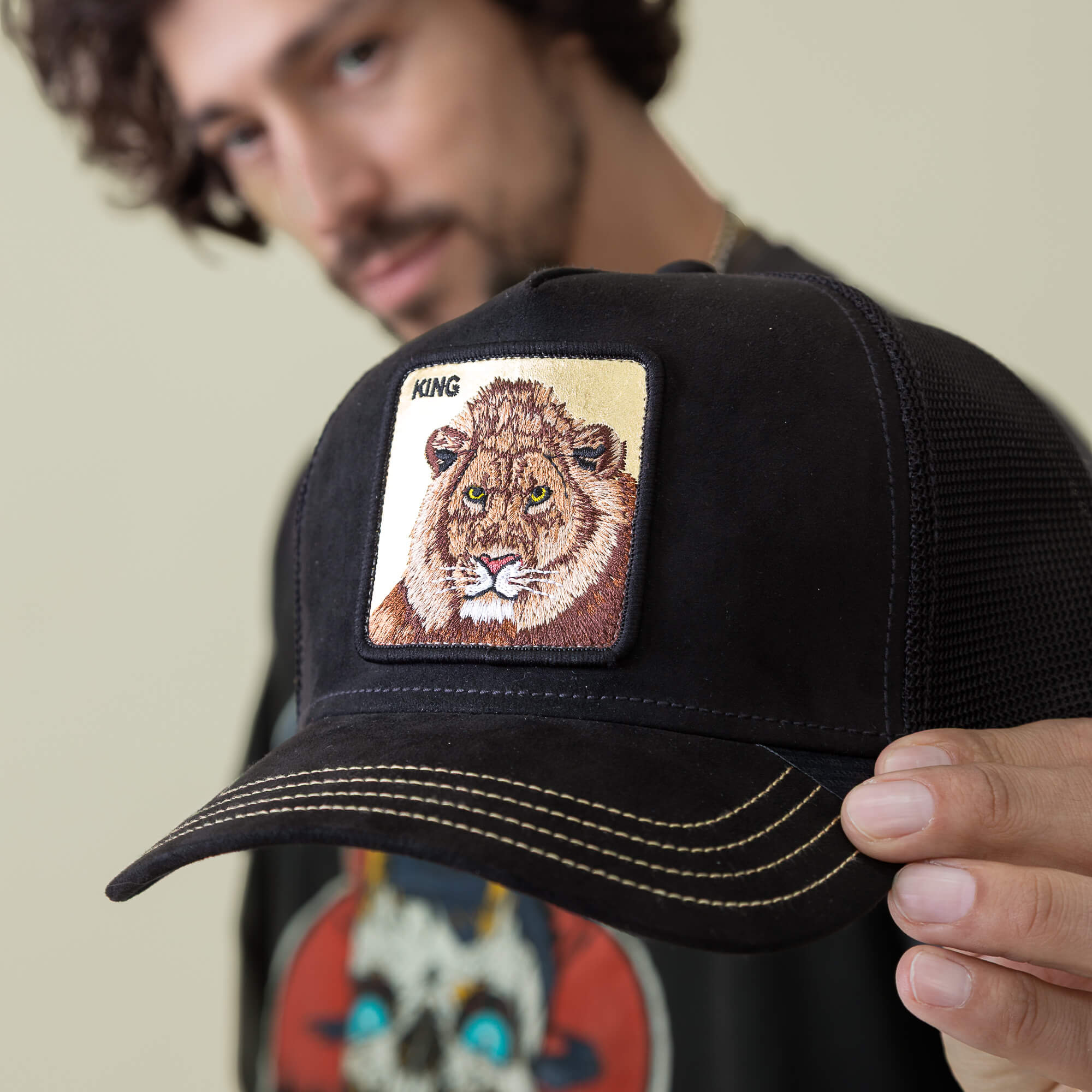 A person holds the Goorin Bros. Golden King cap—a black hat with yellow stitching and a patch of an embroidered Golden Lion and "KING." The wearer’s face appears out of focus in the background.