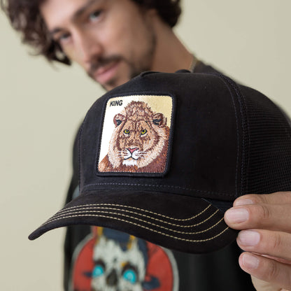 A person holds the Goorin Bros. Golden King cap—a black hat with yellow stitching and a patch of an embroidered Golden Lion and "KING." The wearer’s face appears out of focus in the background.
