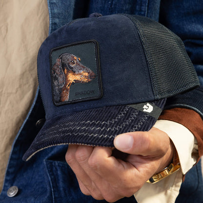 A person holds the Goorin Bros. Shadow Remix, a navy blue mesh trucker hat featuring an embroidered dachshund head patch and the word SHADOW—a true luxury introvert accessory.
