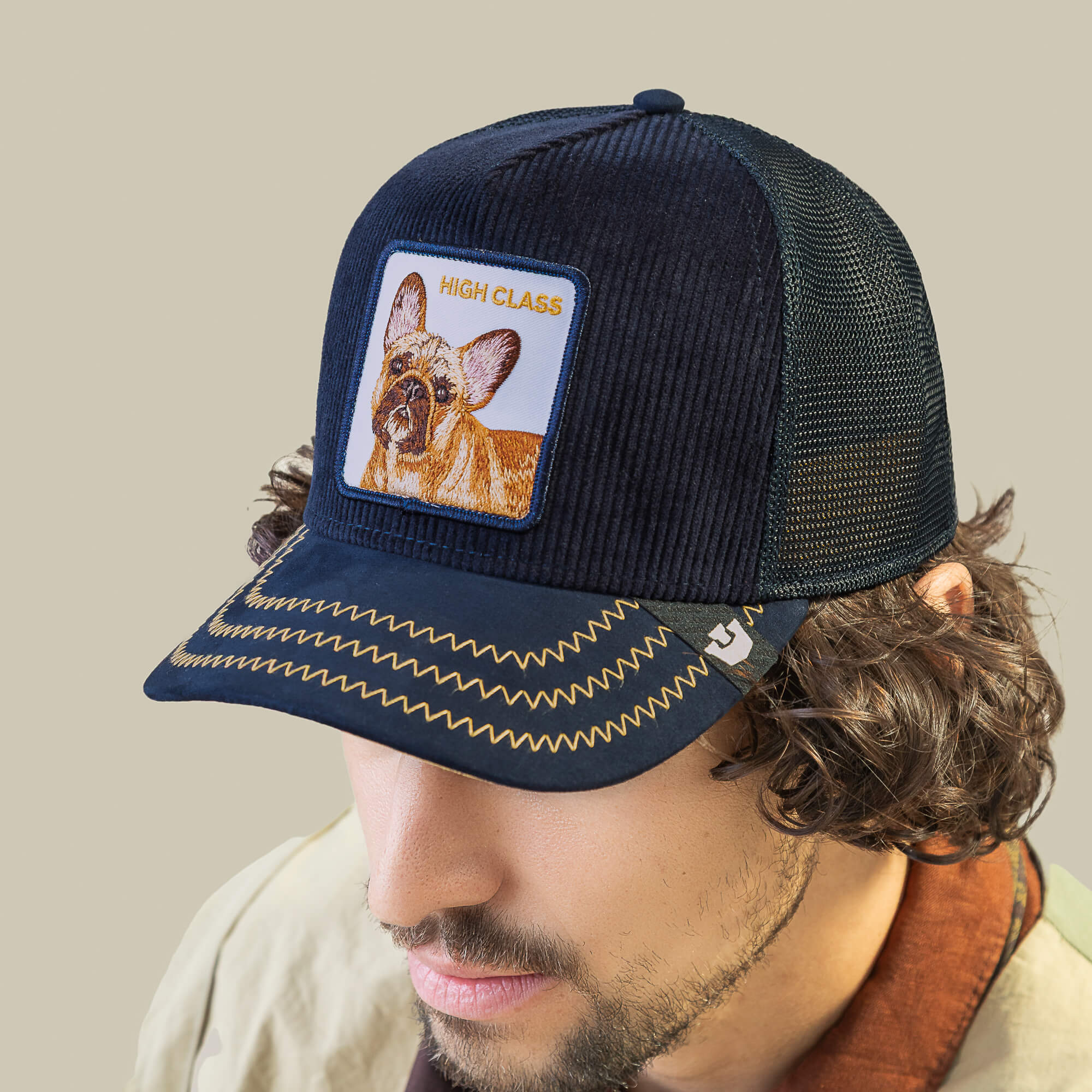 A man wears the Goorin Bros. High Class Remix: a navy corduroy trucker hat with a dog patch, HIGH CLASS text, and yellow zigzag stitching on the brim for unique style.