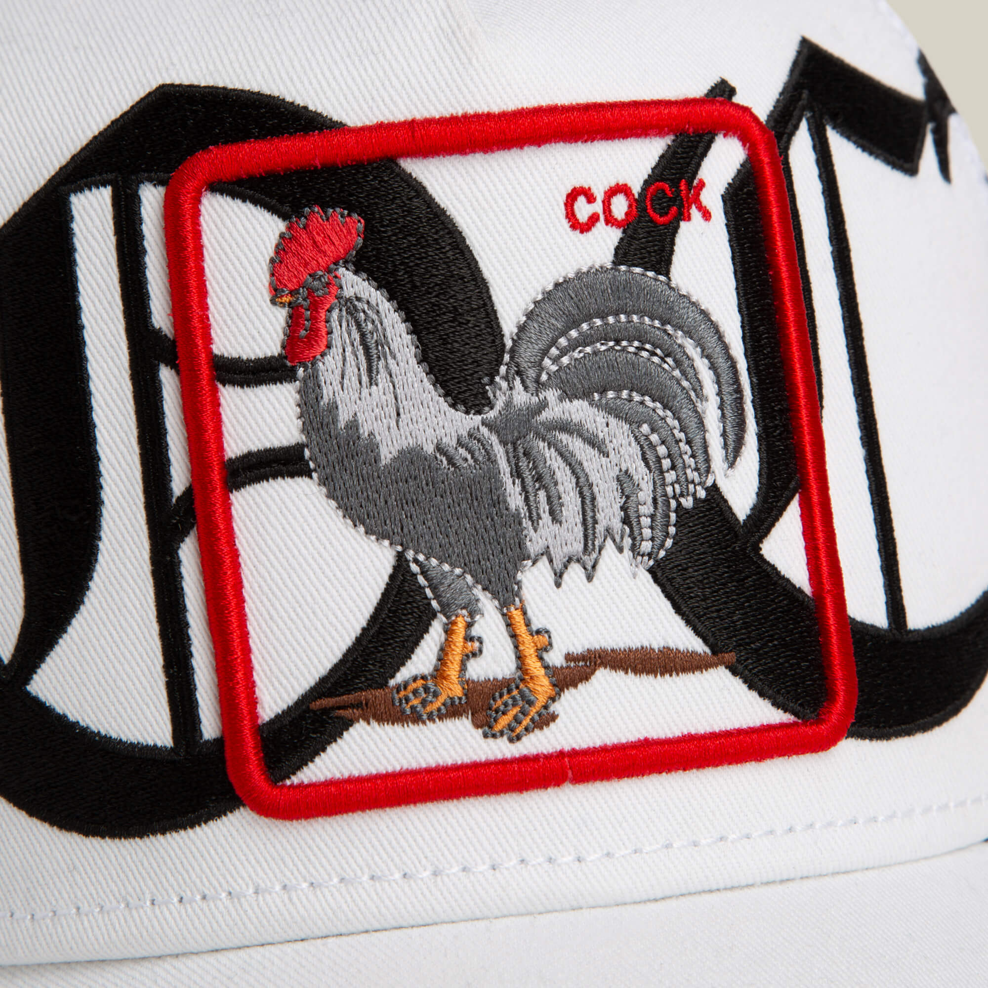 Goorin Bros. Old English Cock hat features a white and black colorway, a rooster patch with bold red "COCK" text, graphic background, and an Old English script accent.