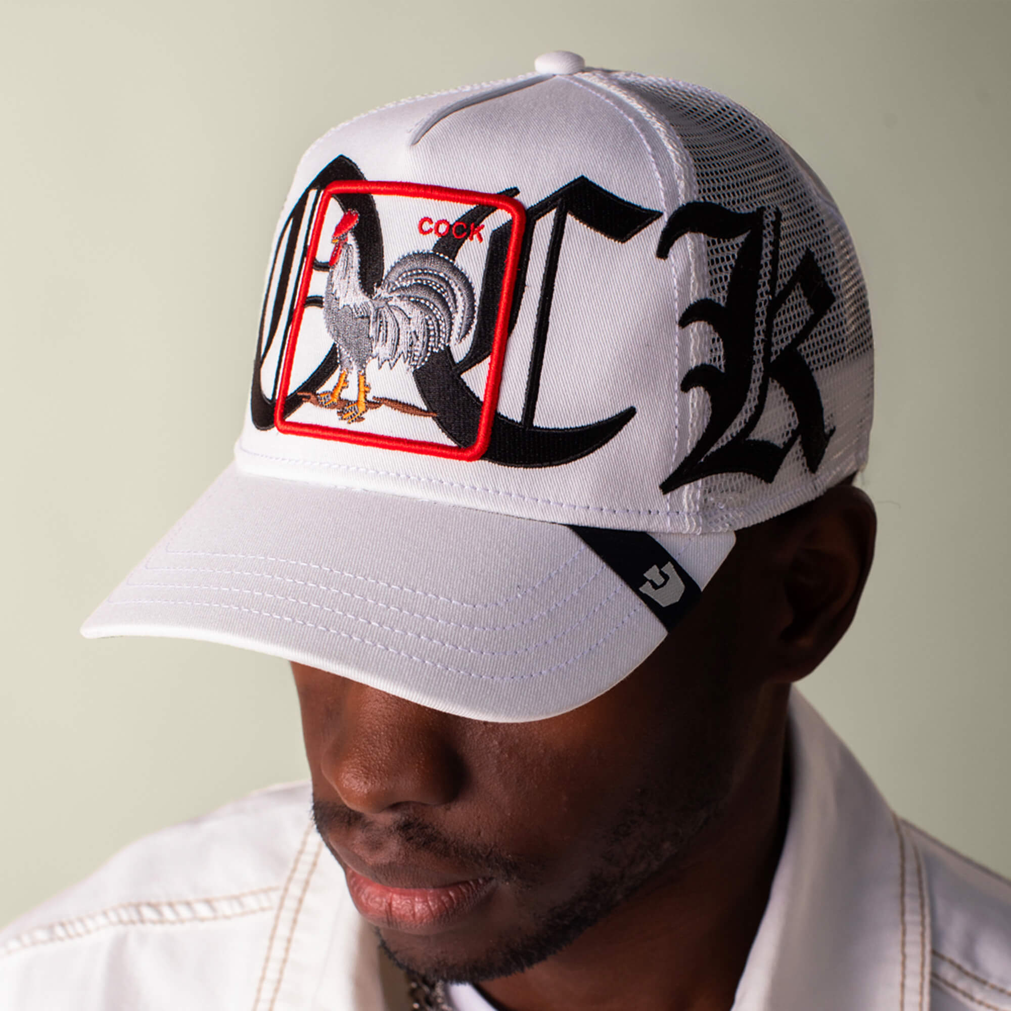 A man wears the Goorin Bros. Old English Cock cap, a white mesh baseball hat featuring a rooster patch and "COCK" embroidered in Old English script on the front, in a classic white and black colorway.