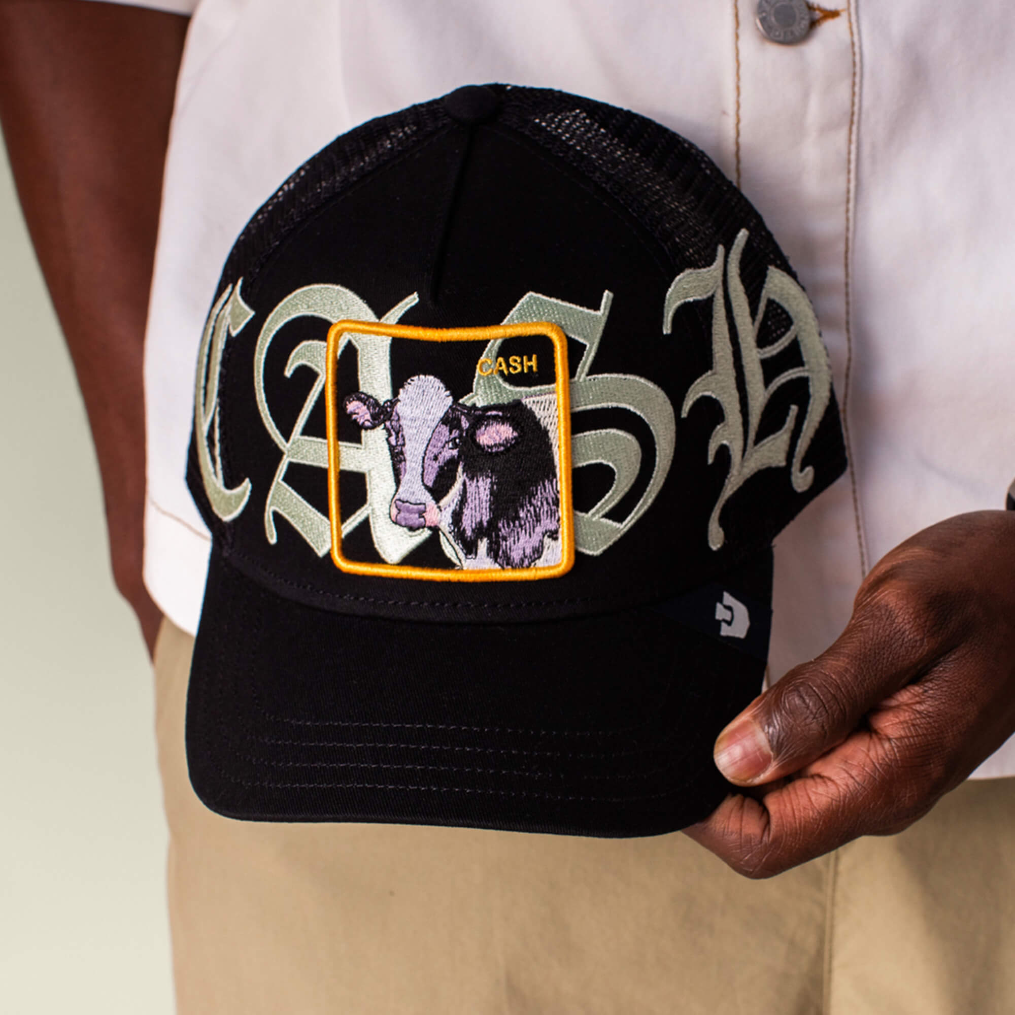 A person holds the Goorin Bros. Old English Cash cap—black, with "CASH" in bold Old English script and a cow patch showing a cow's face and the word CASH, giving a playful farm money vibe.