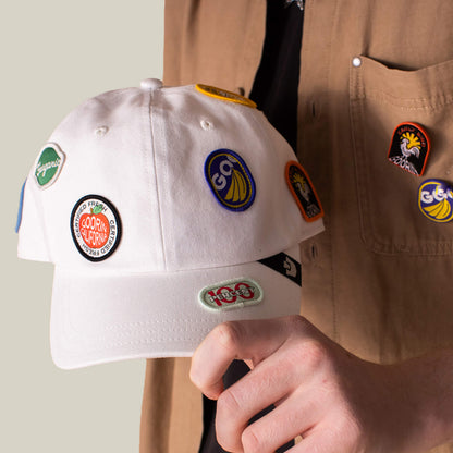A person holds the Goorin Bros. Pop Appeal white canvas baseball cap featuring colorful embroidered patches, with similar badges on their tan shirt pocket.