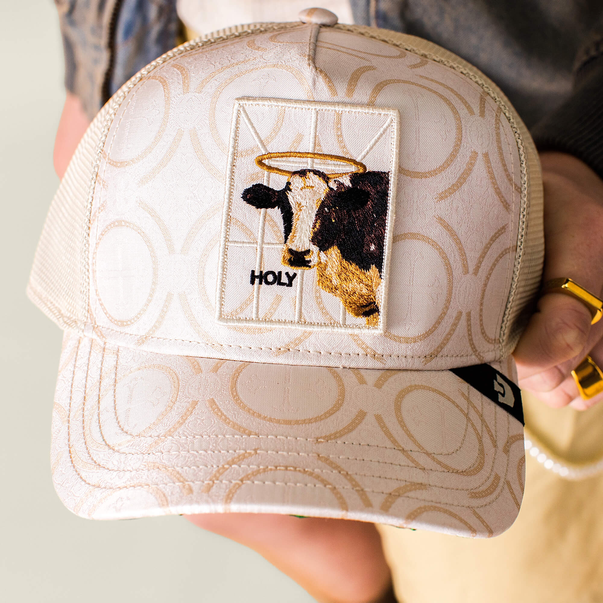 The Religulous - Holy Cow trucker hat by Goorin Bros. features a patterned beige design, a haloed sacred cow patch, and "HOLY" embroidered below, merging faith-inspired imagery with playful humor.