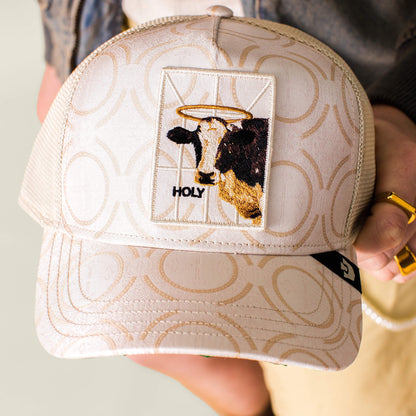 The Religulous - Holy Cow trucker hat by Goorin Bros. features a patterned beige design, a haloed sacred cow patch, and "HOLY" embroidered below, merging faith-inspired imagery with playful humor.