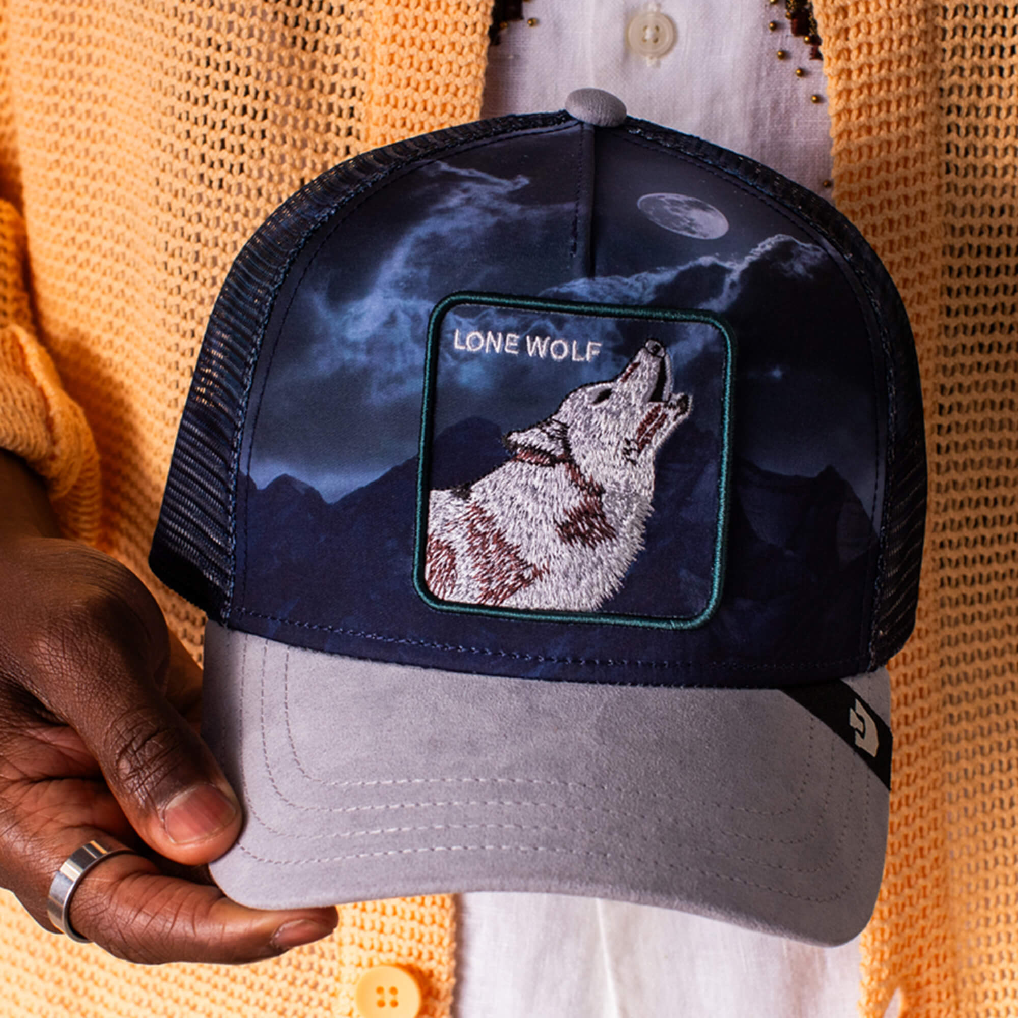 A person holds a Goorin Bros. "Wolf In The Element" trucker hat, which features a patch of a howling wolf with a moon and mountains. They are wearing a yellow cardigan over a white shirt.