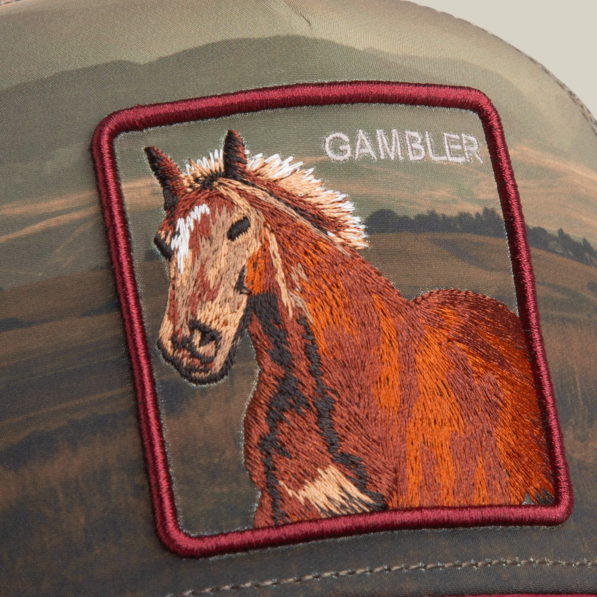 A close-up of the Goorin Bros. Stallion In The Element trucker hat, showcasing an embroidered brown horse patch with "GAMBLER" above it on an earth-toned landscape backdrop.