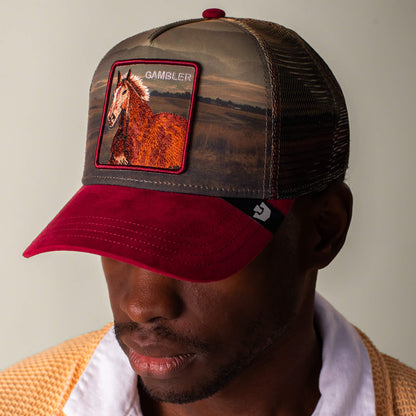 A man wearing a Goorin Bros. Stallion In The Element trucker hat featuring a red brim and a patch with a horse and the word GAMBLER.