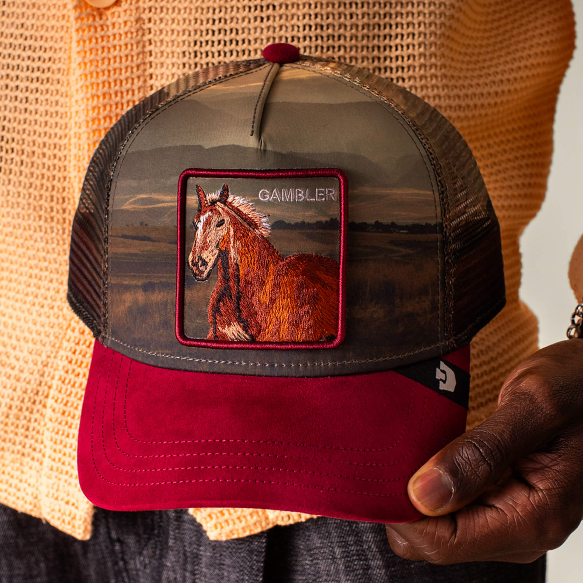 A person holds a Goorin Bros. Stallion In The Element trucker hat with a red brim and a patch featuring a horse and the word GAMBLER on the front.