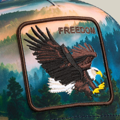 A close-up of the Goorin Bros. "Eagle In The Element" hat features a patch with an embroidered bald eagle in flight and the word FREEDOM above it, set against a scenic forest landscape background.