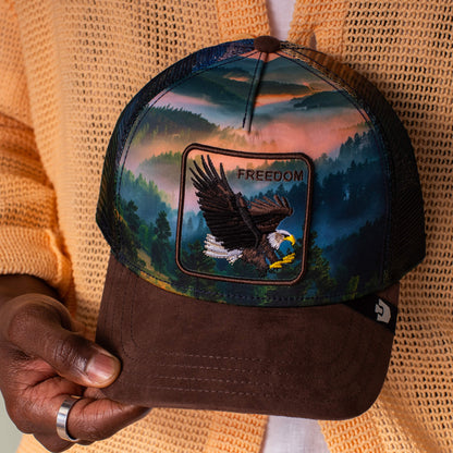A person holds the Goorin Bros. "Eagle In The Element" hat, featuring an earth-toned landscape and a patch with a flying bald eagle and "FREEDOM," set against a scenic forest and mountain backdrop.
