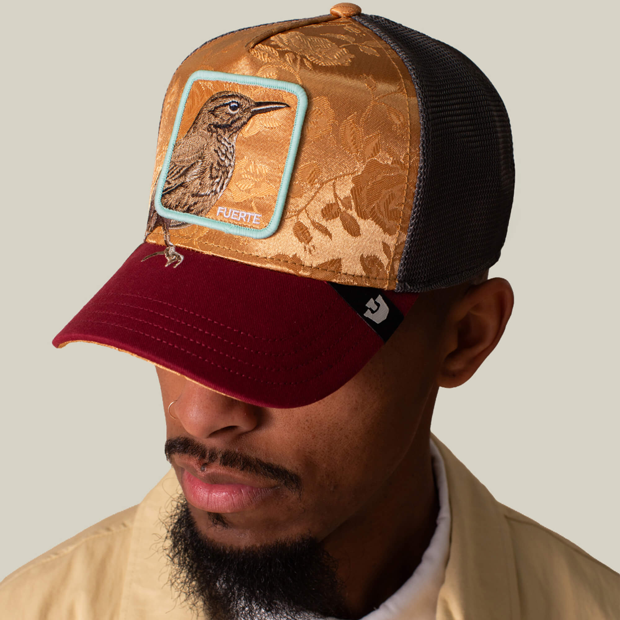 A man wearing the Goorin Bros. Costa Rica Thrush gold and burgundy trucker hat with a charcoal mesh back and a bird patch labeled FUERTE on the front, looking slightly downward.