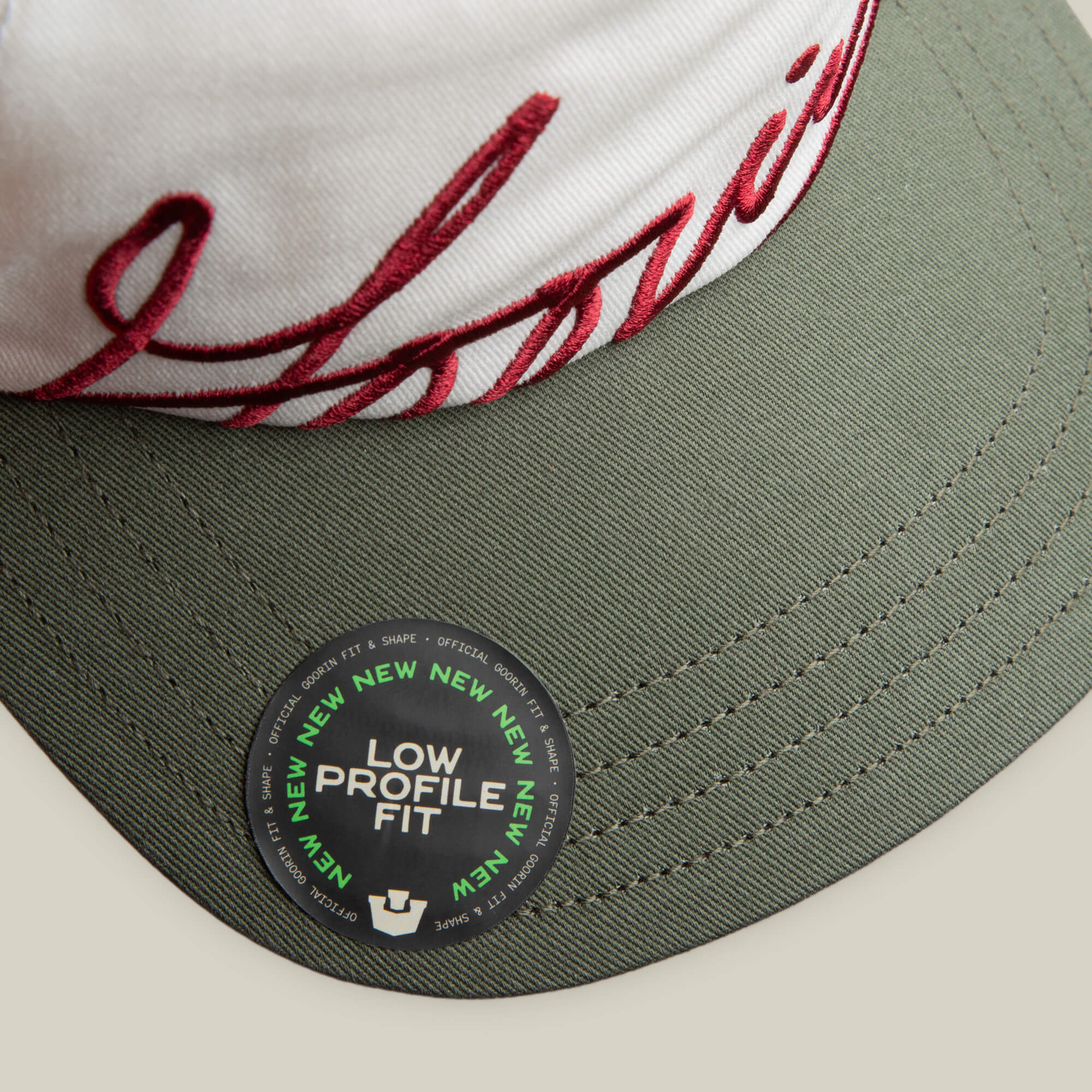 A close-up of the Goorin Bros. "Goorin Script" white and green cap with white and red embroidery, plus a sticker reading LOW PROFILE FIT with NEW repeated around the edge.