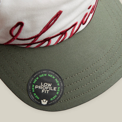 A close-up of the Goorin Bros. "Goorin Script" white and green cap with white and red embroidery, plus a sticker reading LOW PROFILE FIT with NEW repeated around the edge.