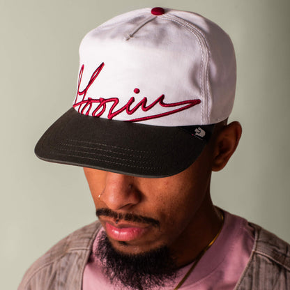 A man wearing the Goorin Bros. Goorin Script white and black full fabric baseball cap with red script on the front, along with a pink shirt and a light brown jacket.