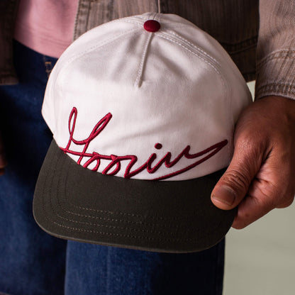 A person holds a white and green Goorin Bros. cap with a dark brim and "Goorin Script" embroidered in bold on the front.