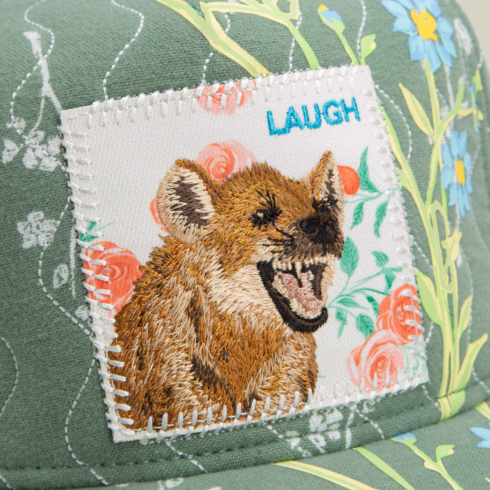 The Goorin Bros. Vol. 1 - Quiltbag: Laugh hat is a green floral trucker cap with a stitched patch of a laughing hyena and "LAUGH" embroidered above, blending playful style with unique patchwork patterns.