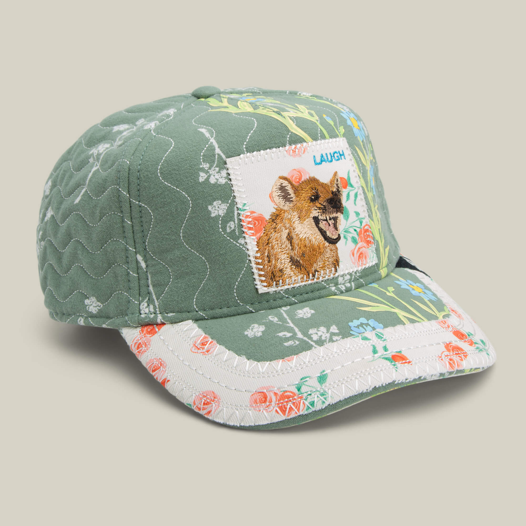 The Goorin Bros. Vol. 1 - Quiltbag: Laugh trucker hat features green and white floral patchwork, a cheerful hyena patch, and “LAUGH” embroidery—bringing playful charm to your look.