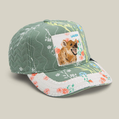The Goorin Bros. Vol. 1 - Quiltbag: Laugh trucker hat features green and white floral patchwork, a cheerful hyena patch, and “LAUGH” embroidery—bringing playful charm to your look.
