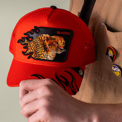 A person wears the red Blazing Flames trucker cap by Goorin Bros., featuring an embroidered BLAZING patch and black flame accents, paired with a tan shirt decorated with colorful pins.