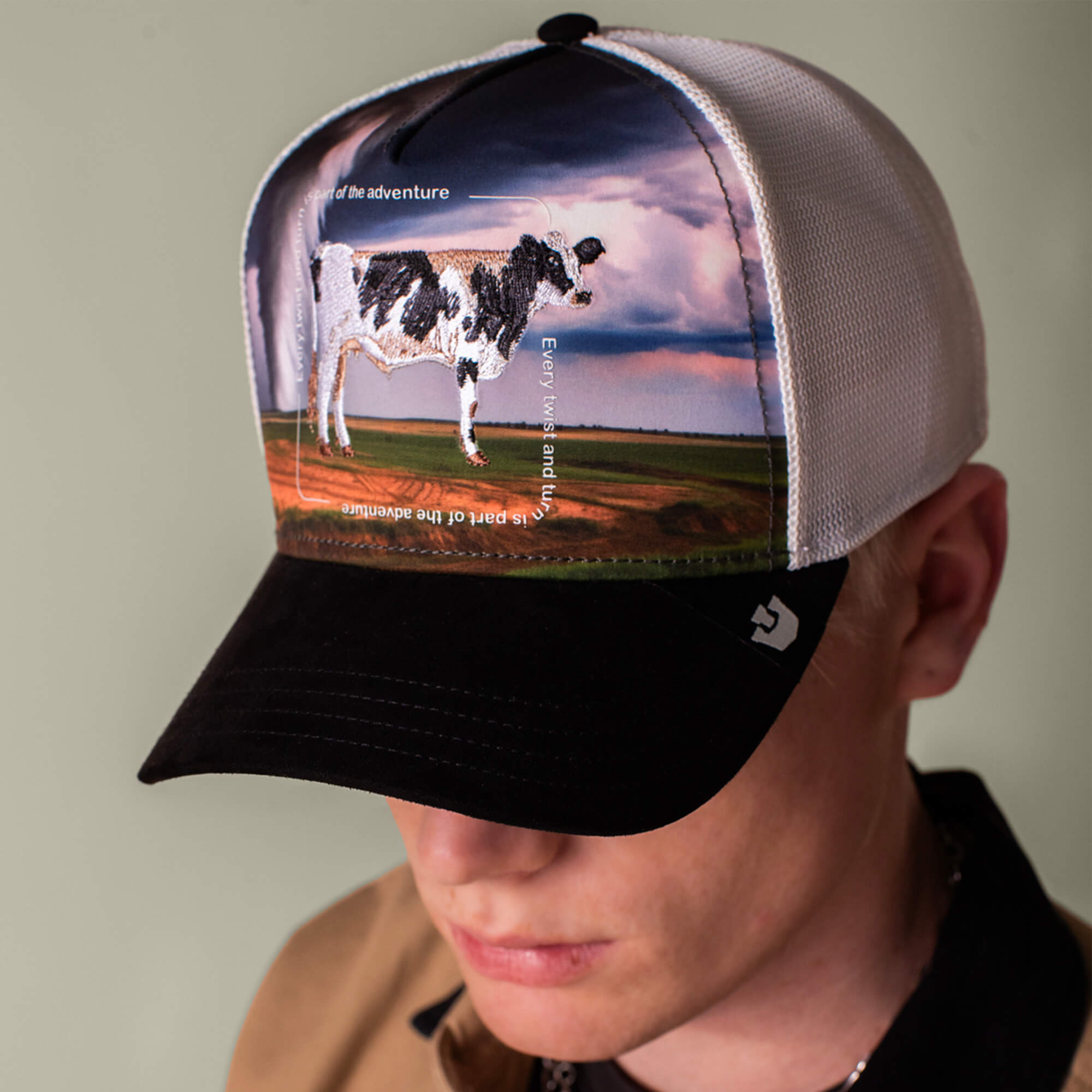 A person wears the Goorin Bros. "Through the Storm - Cow" black and white trucker cap with a cow patch, featuring the words "Enjoy the adventure," set against a scenic landscape backdrop.