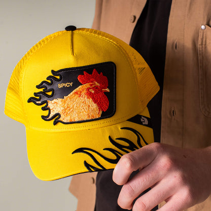 A person holds the Goorin Bros. Spicy Flames trucker hat, a bright yellow cap featuring an embroidered rooster patch, the word "SPICY," and striking black flame designs.