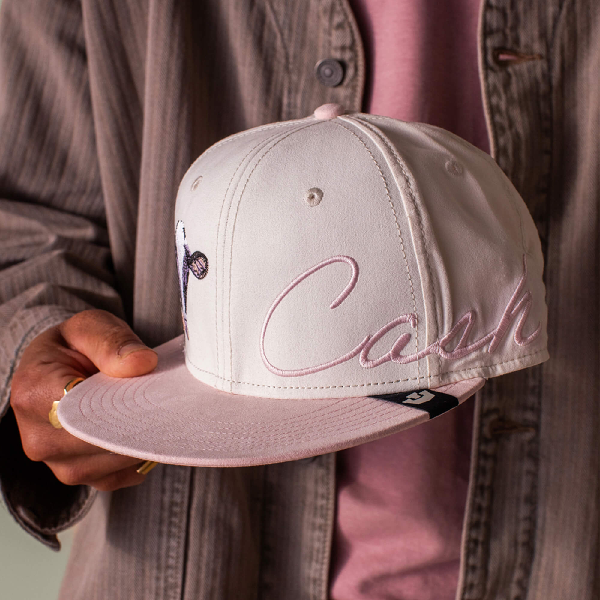 Someone holds the Goorin Bros. 6-Panel Cash Script hat in light pink and white cow print, featuring “Cash” embroidered on the side. They are dressed in a pink shirt and brown jacket.