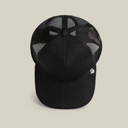 The Clean Denim Trucker by Goorin Bros. is a black mesh trucker hat with a solid front panel and curved brim, made in the USA. A small side tag with a white logo adds a classic touch. Shown from above on a neutral background.
