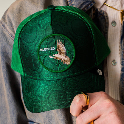 A person wearing the Goorin Bros. "Religulous - Blessed" deep green hat with a textured pattern and a flying bird patch, holding the cap's brim with one hand.