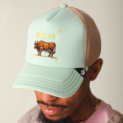 A person wears the Neo Farm - Bully Bull mesh trucker hat by Goorin Bros., in light green and beige with an embroidered brown cow and "BULLY" on the front, paired with calm collected apparel for a structured yet flexible look.