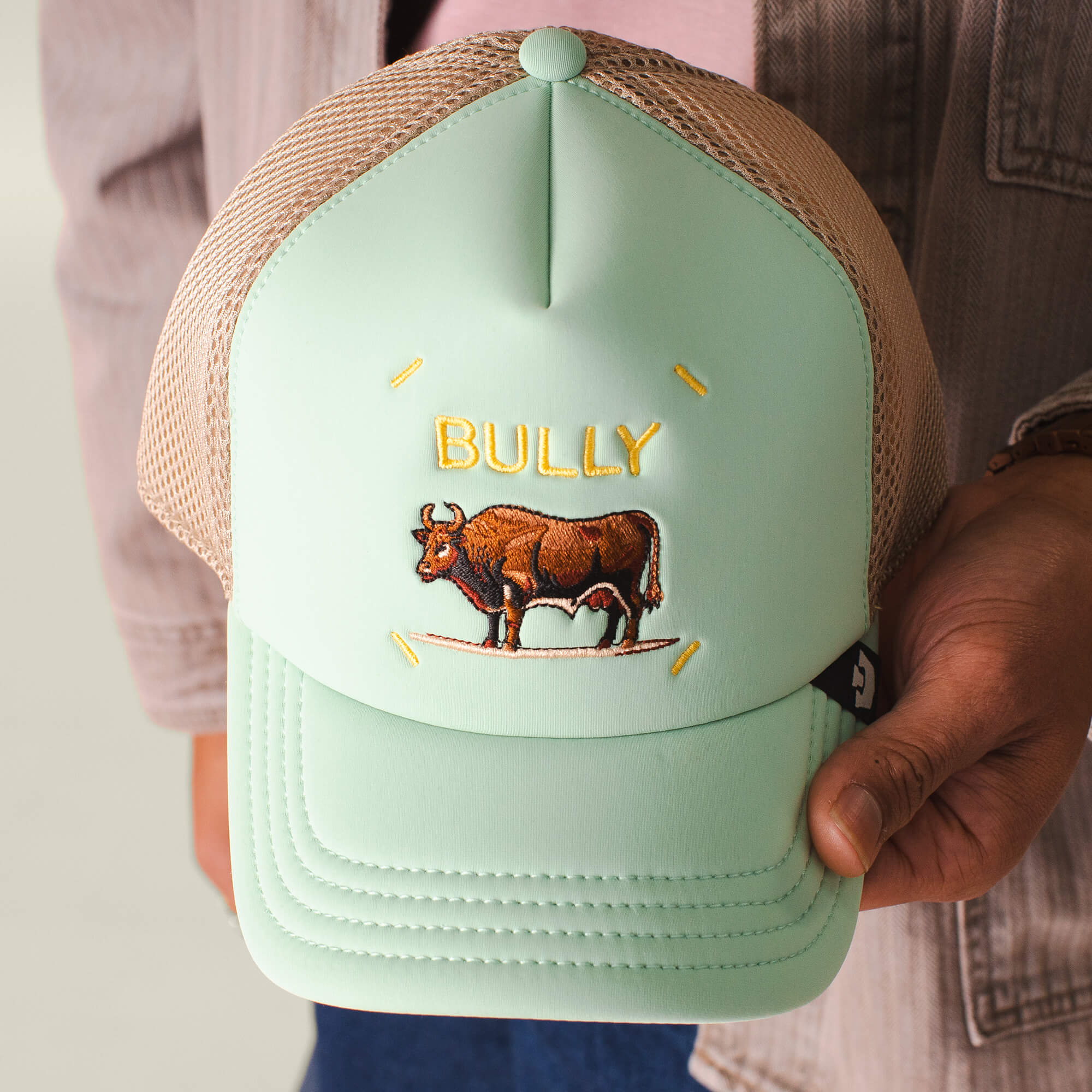 A person holds the Neo Farm - Bully Bull trucker hat by Goorin Bros., featuring a mint green color, a cow patch, and "BULLY" embroidered on the front, highlighting its structured flexible fit and calm collected style.