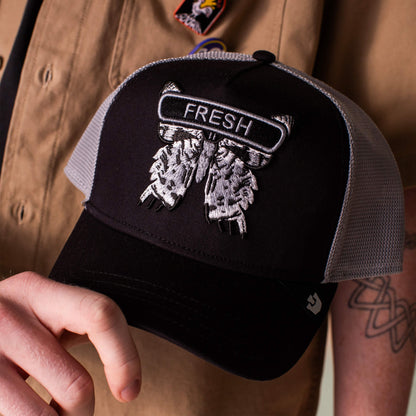 A person in a brown shirt holds the Goorin Bros. Classic Rocker - Fresh hat, a black and grey mesh cap with embroidered feathers and "FRESH" on the front, highlighting its bold rocker style.