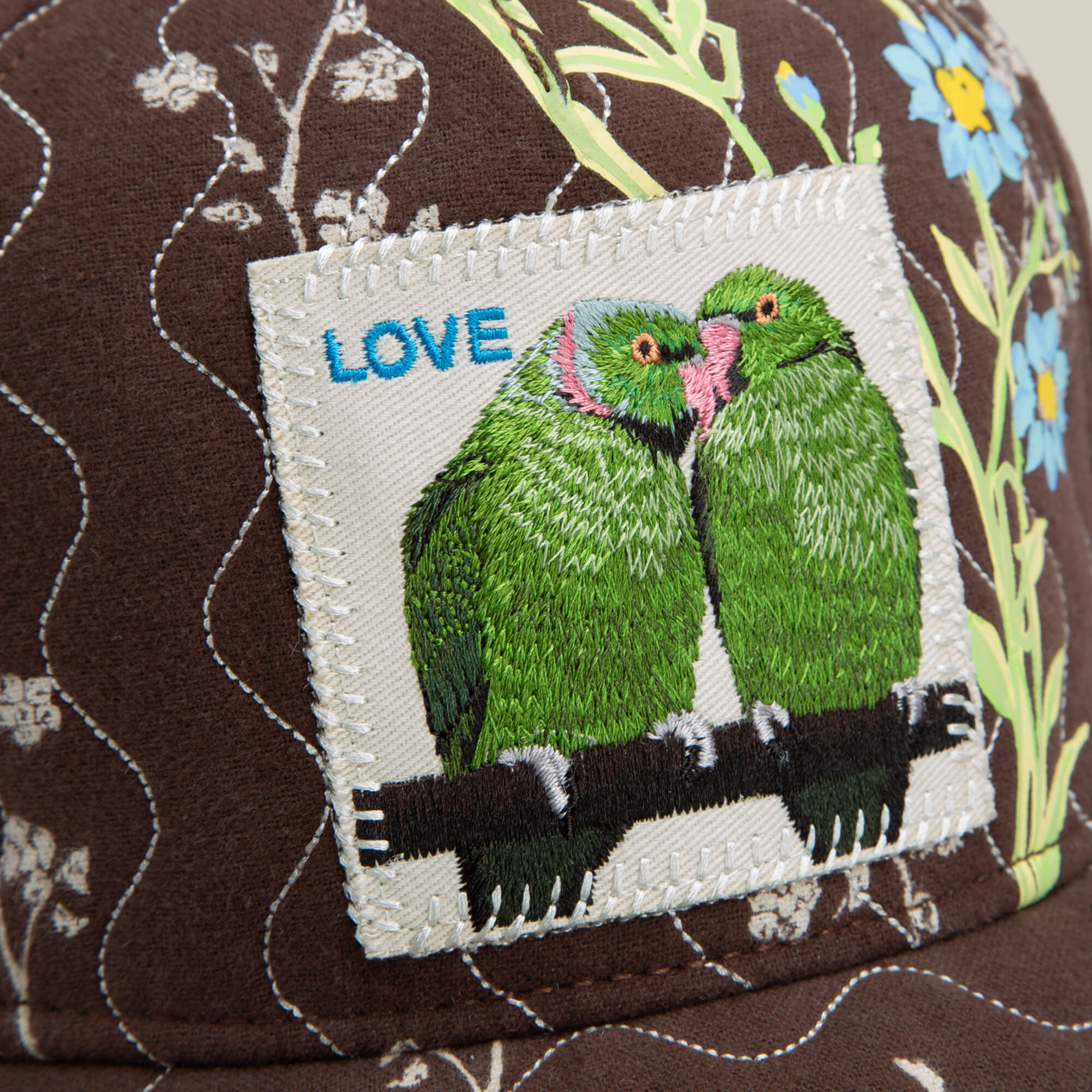 Close-up of the Goorin Bros. Vol. 1 - Quiltbag: Love OG Trucker hat in chocolate brown quilted fabric, with a hand-stitched patch showing two green parrots touching beaks, the word LOVE, and blue flowers in the background.