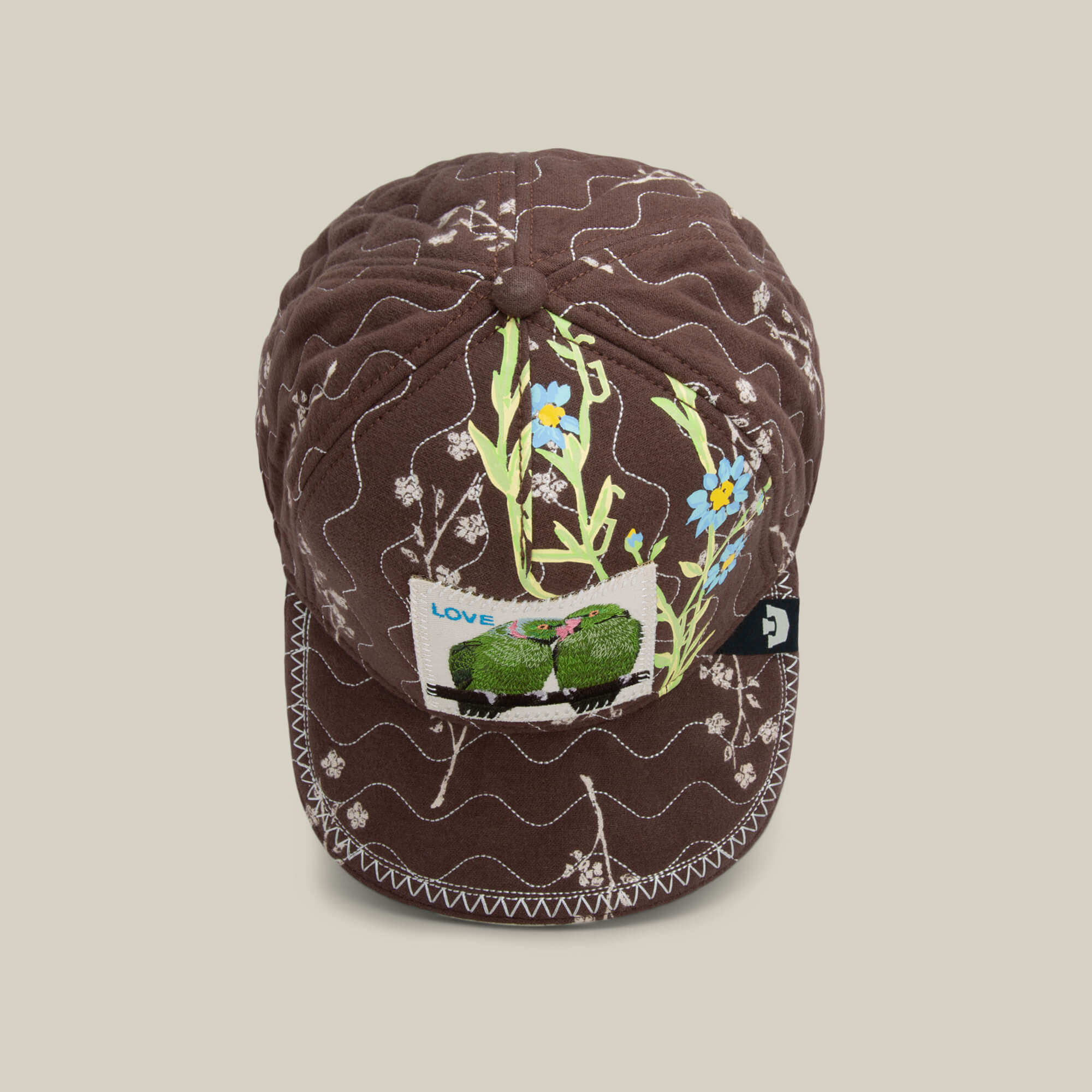 The Goorin Bros. Vol. 1 - Quiltbag: Love is a hand-stitched OG Trucker cap in chocolate brown quilting with white stitching, yellow and blue floral embroidery, and a “LOVE” patch featuring two green frogs.