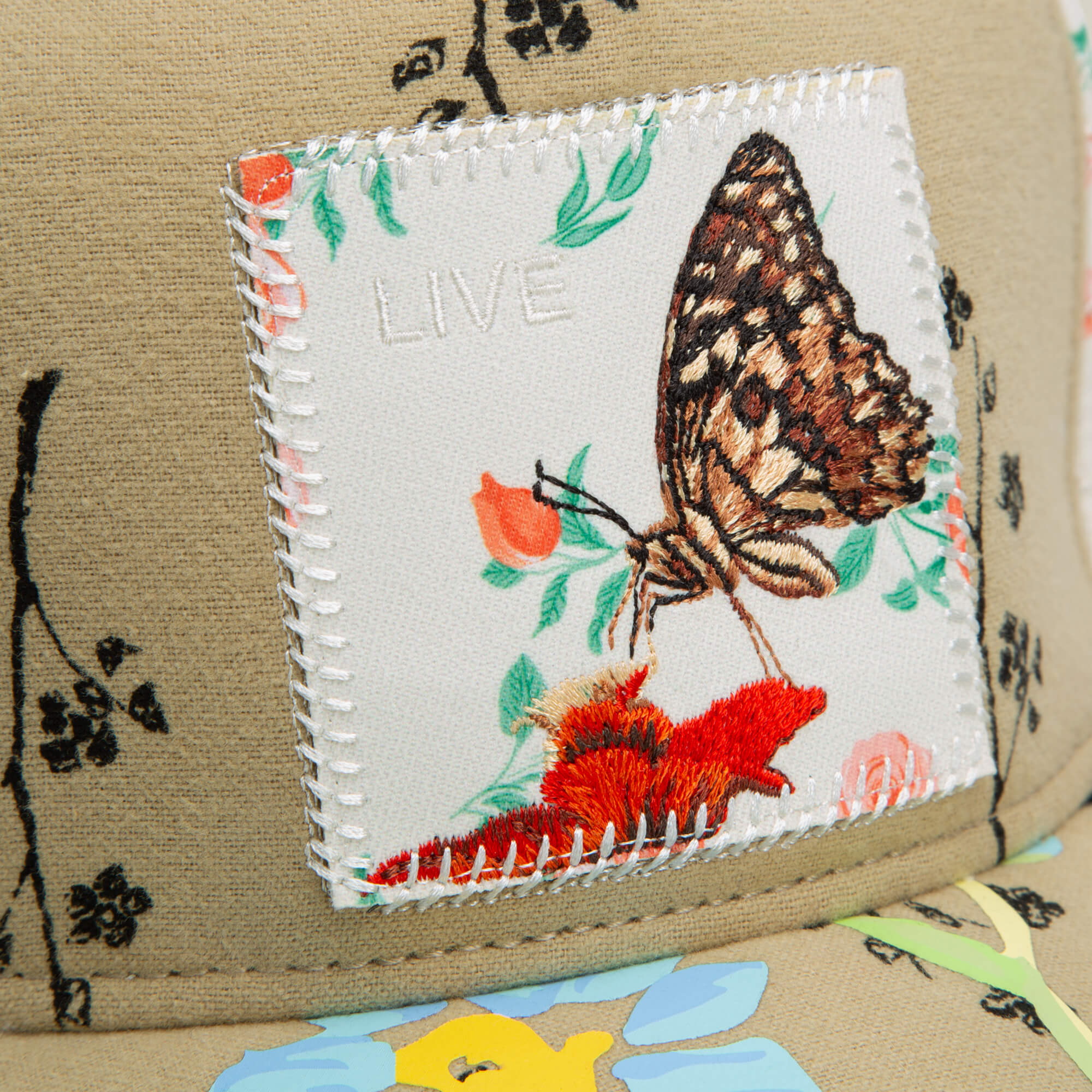 Close-up of a fabric patch stitched on tan, with a butterfly and floral embroidery plus "LIVE" above it, from the Vol. 1 – Quiltbag: Live by Goorin Bros., set against bold black and colorful patterns reminiscent of OG Trucker hat style.