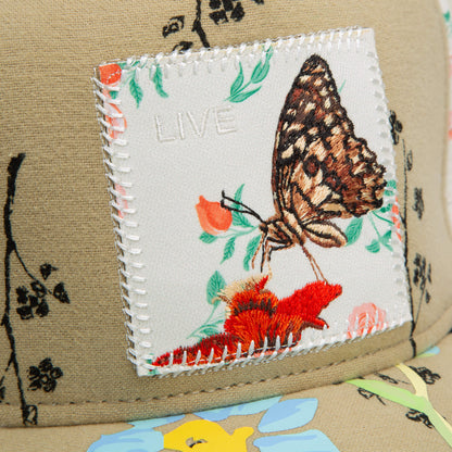 Close-up of a fabric patch stitched on tan, with a butterfly and floral embroidery plus "LIVE" above it, from the Vol. 1 – Quiltbag: Live by Goorin Bros., set against bold black and colorful patterns reminiscent of OG Trucker hat style.