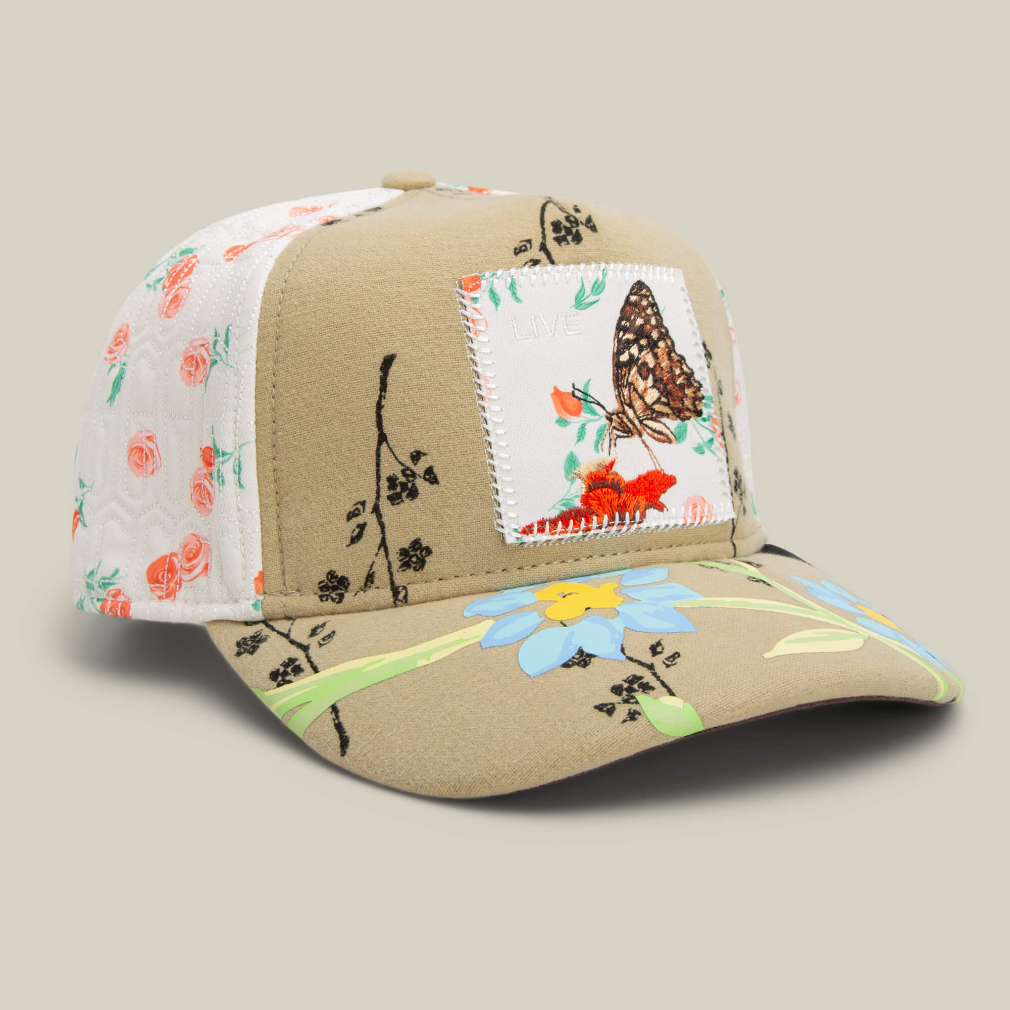 The Goorin Bros. Vol. 1 - Quiltbag: Live OG Trucker hat features tan and white panels, a butterfly design patch on the front, and blue and red floral embroidery with green stems on the brim and panels.