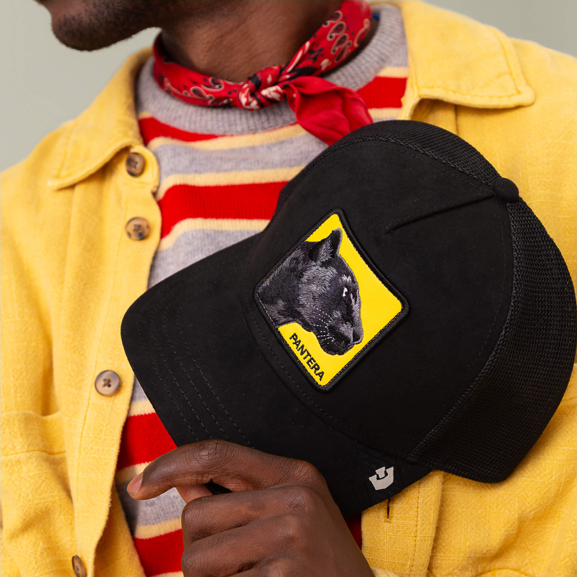 A person in a yellow jacket and red striped shirt holds a Goorin Bros. Pantera Gialla black cap with a panther graphic and the word PANTERA, paired with Pantera Gialla agile sneakers in luxe microsuede.